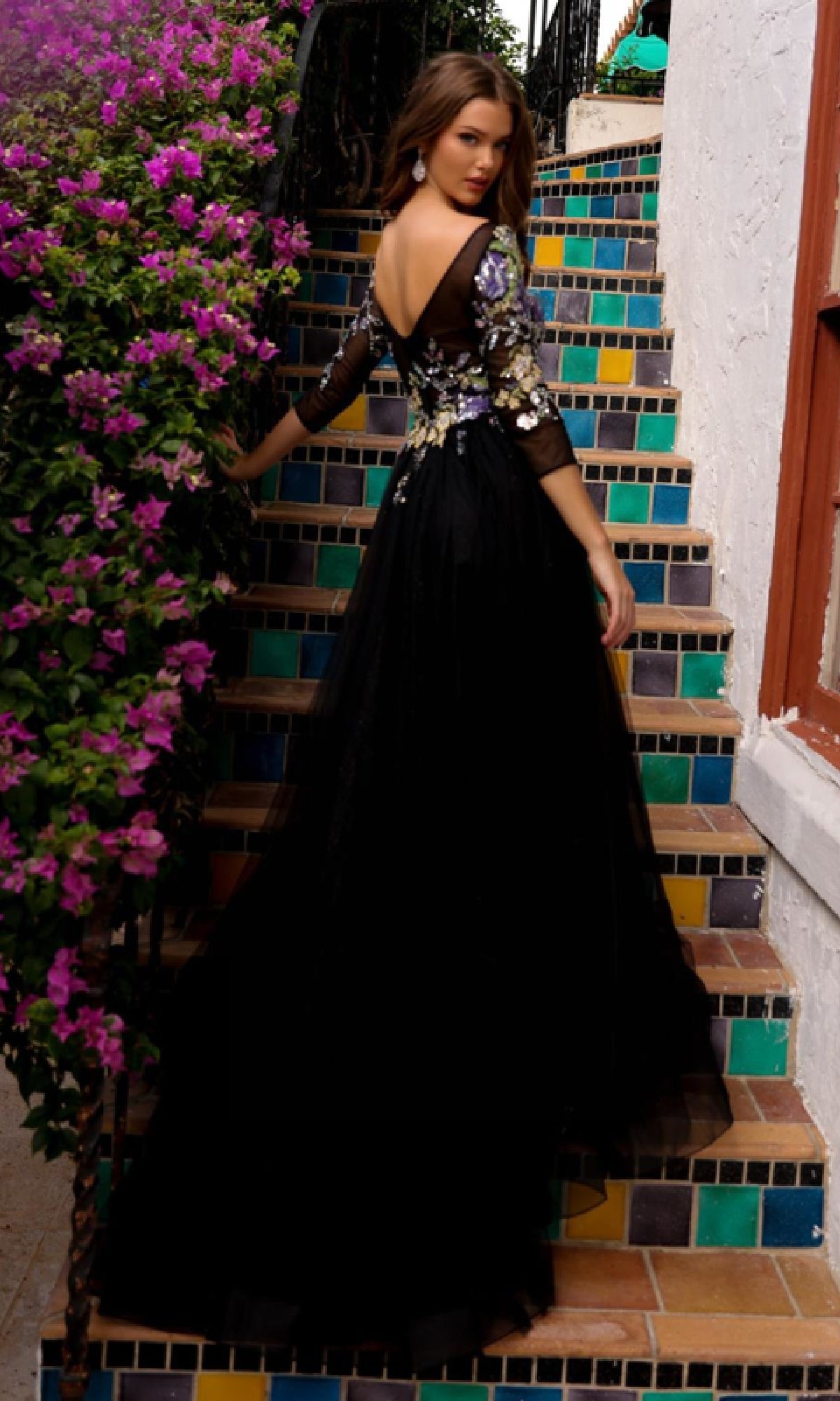 A woman in the Long Black Formal Dress: Amelia Couture TM1021, featuring floral details, stands on colorful tiled stairs beside blooming purple flowers, glancing back over her shoulder.